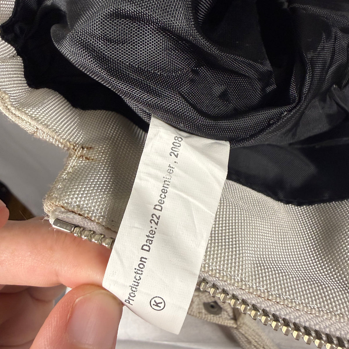 A close-up of a garment's interior showing a white paper tag held by a hand, with a black mesh lining, beige fabric, and a silver zipper visible.