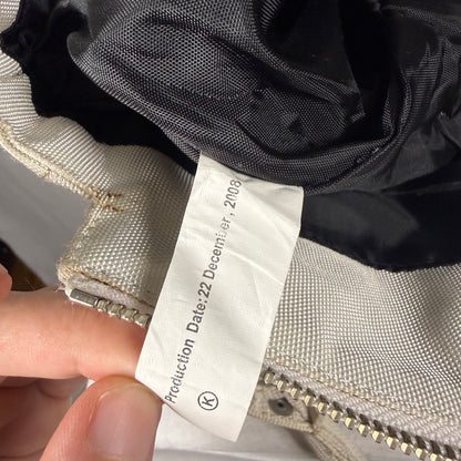 A close-up of a garment's interior showing a white paper tag held by a hand, with a black mesh lining, beige fabric, and a silver zipper visible.