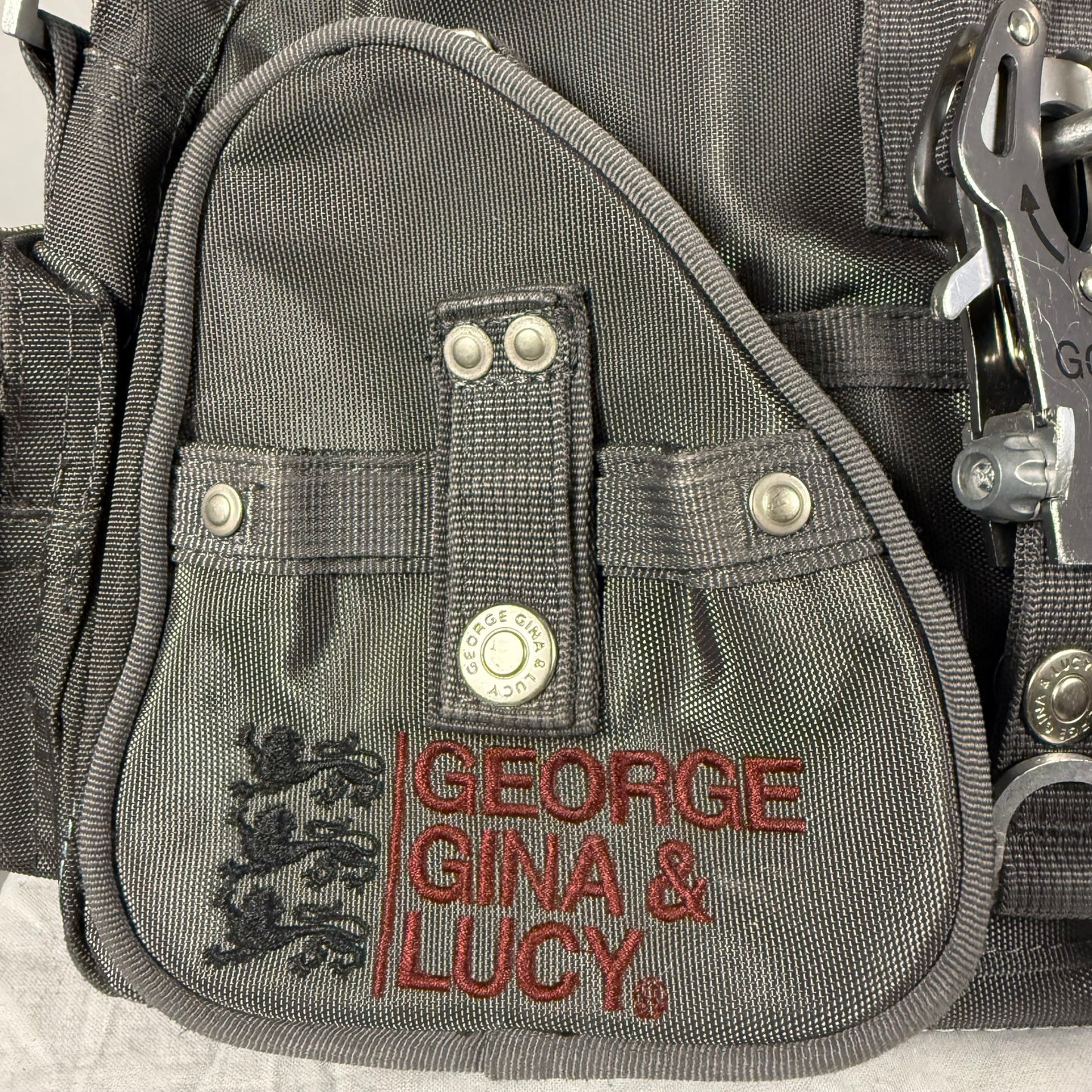 Close-up of a gray nylon utility pouch with red embroidered text reading 'GEORGE GINA & LUCY' and a black crest logo, plus silver metal snaps and clips.