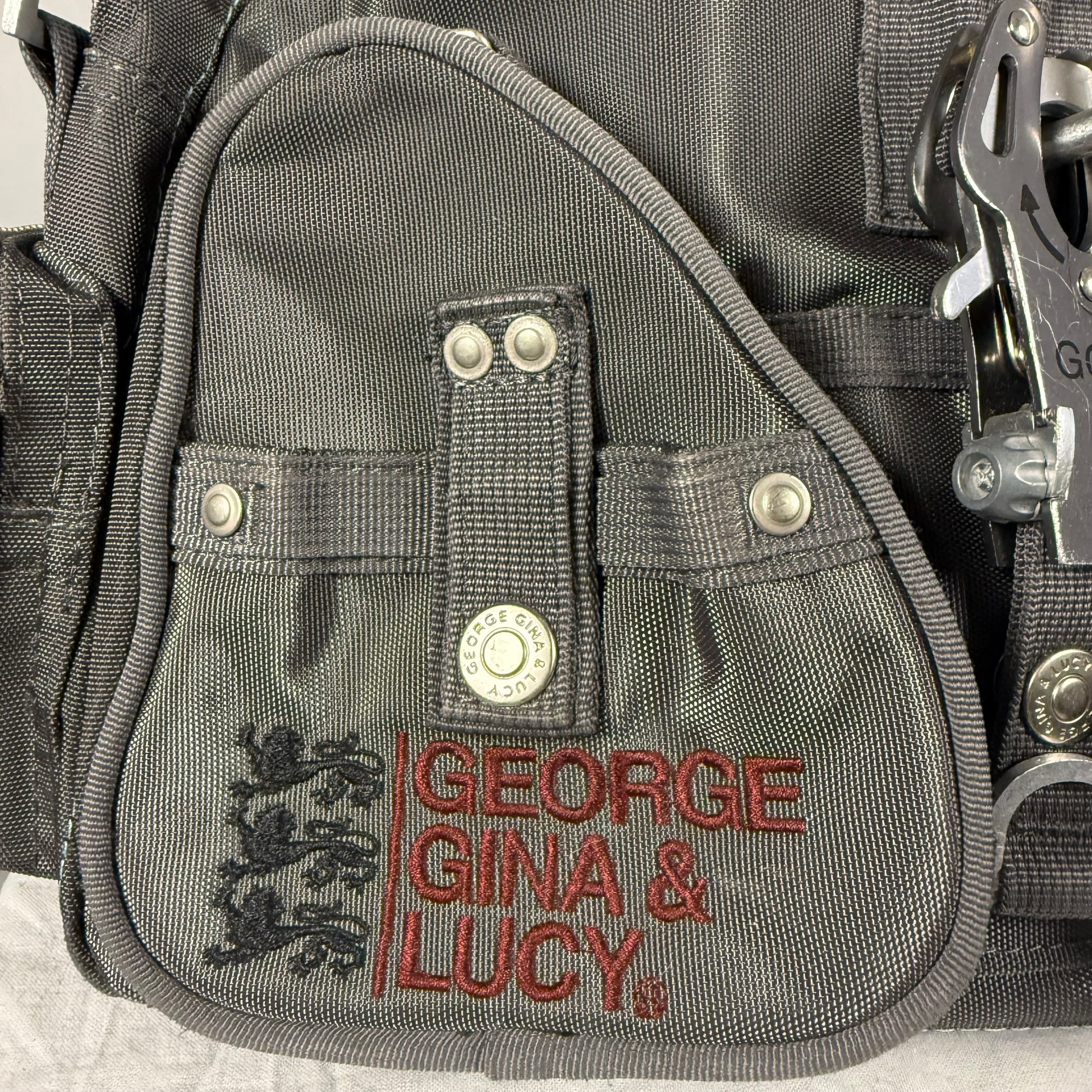 Close-up of a gray nylon utility pouch with red embroidered text reading 'GEORGE GINA & LUCY' and a black crest logo, plus silver metal snaps and clips.