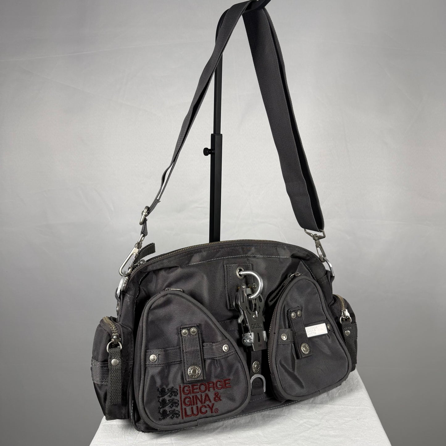 A black multi-pocket crossbody bag with a long shoulder strap, displayed on a white surface against a gray background. The front pocket features the text 'GEORGE GINA & LUCY' in red.