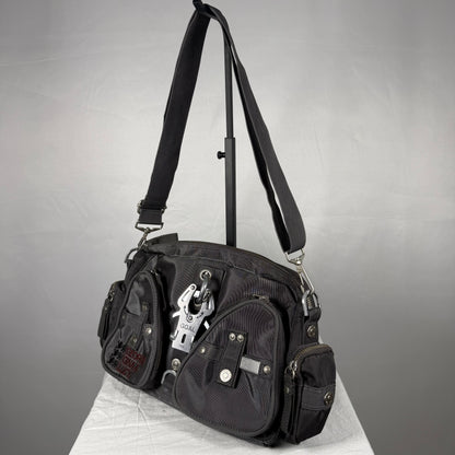 A black canvas shoulder bag with multiple utility pockets and silver hardware, displayed on a white surface against a gray studio background.