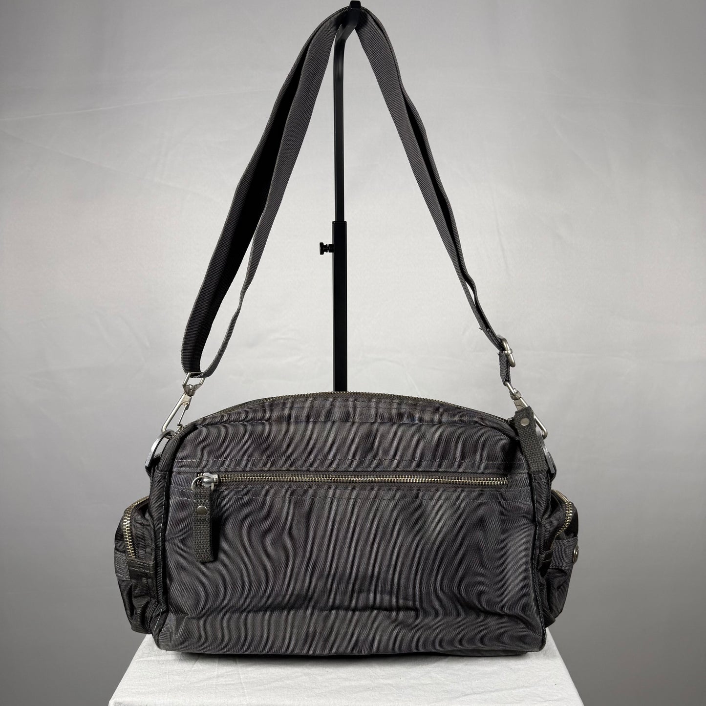 A black nylon shoulder bag with a front zippered pocket and side pockets, displayed on a white surface with a black stand against a gray background.