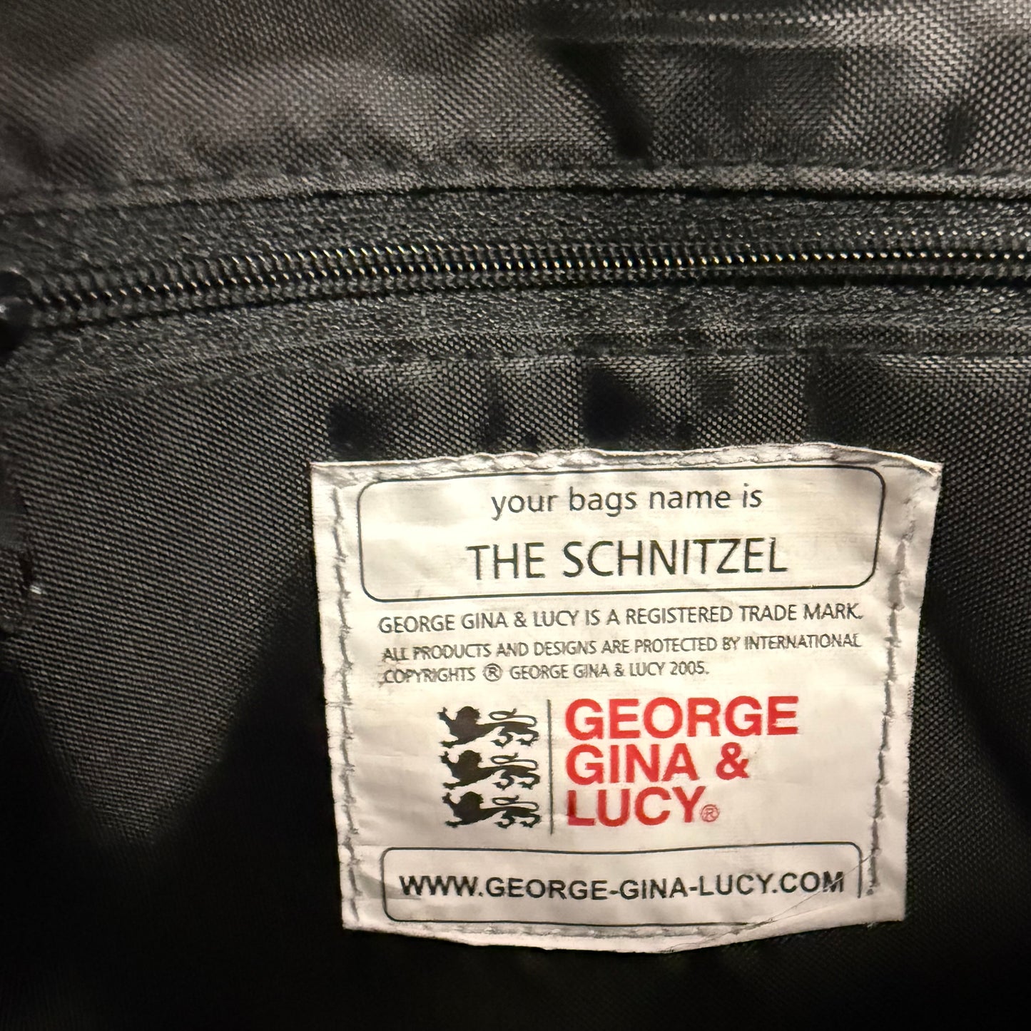 A close-up of a white sewn-in label on a black fabric bag, featuring the text 'THE SCHNITZEL' and the George Gina & Lucy brand name in red and black.