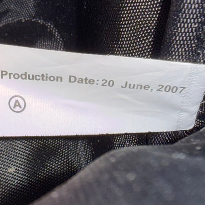 Close-up of a white fabric label sewn onto black textured mesh fabric, showing the printed text 'Production Date: 20 June, 2007' and a circular letter 'A'.
