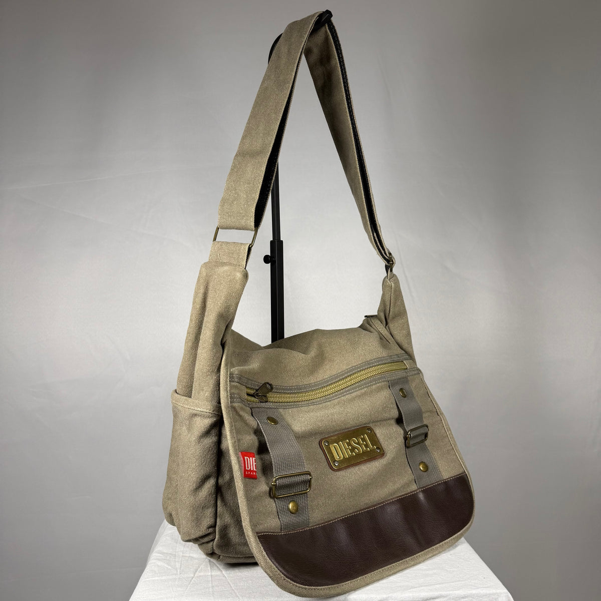 A Diesel canvas shoulder bag in beige and brown, featuring a front zippered pocket with a brown leather patch, side pockets, and a wide adjustable shoulder strap, displayed on a white surface against a gray background.