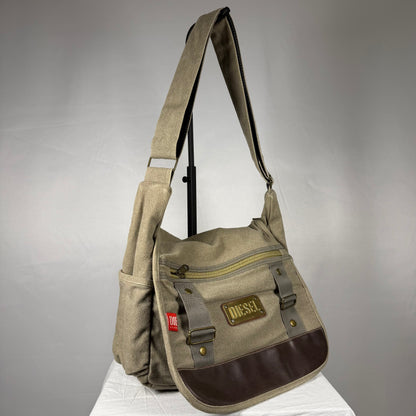 A Diesel canvas shoulder bag in beige and brown, featuring a front zippered pocket with a brown leather patch, side pockets, and a wide adjustable shoulder strap, displayed on a white surface against a gray background.