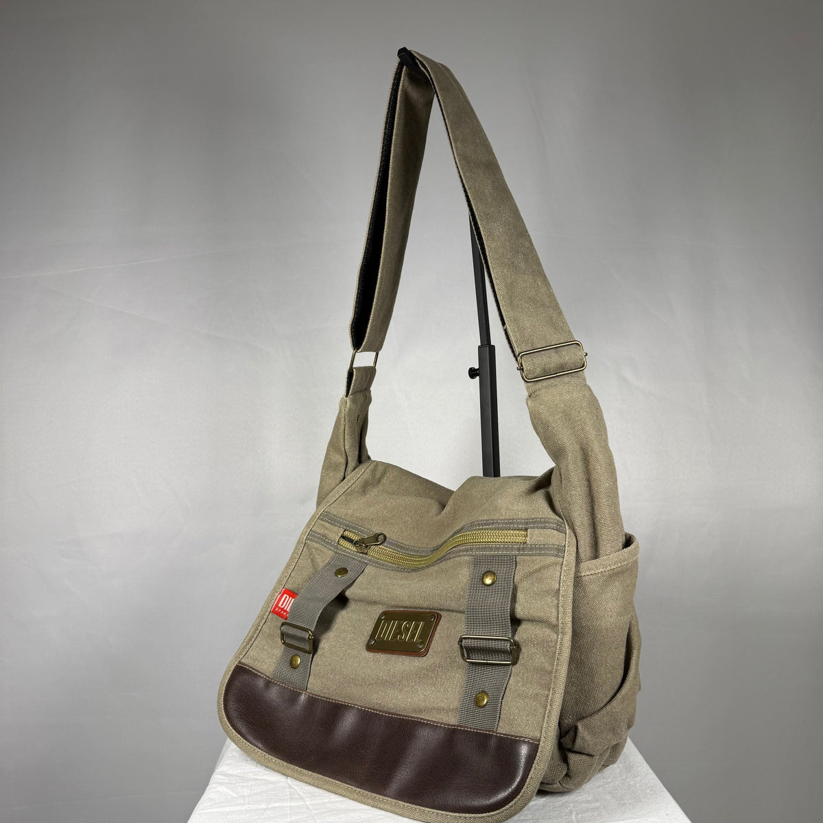 A canvas shoulder bag in olive green with dark brown leather accents and brass hardware, displayed on a white surface against a gray background.