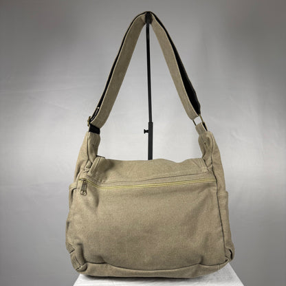 A beige canvas shoulder bag with a wide adjustable strap, displayed on a stand against a gray studio background.