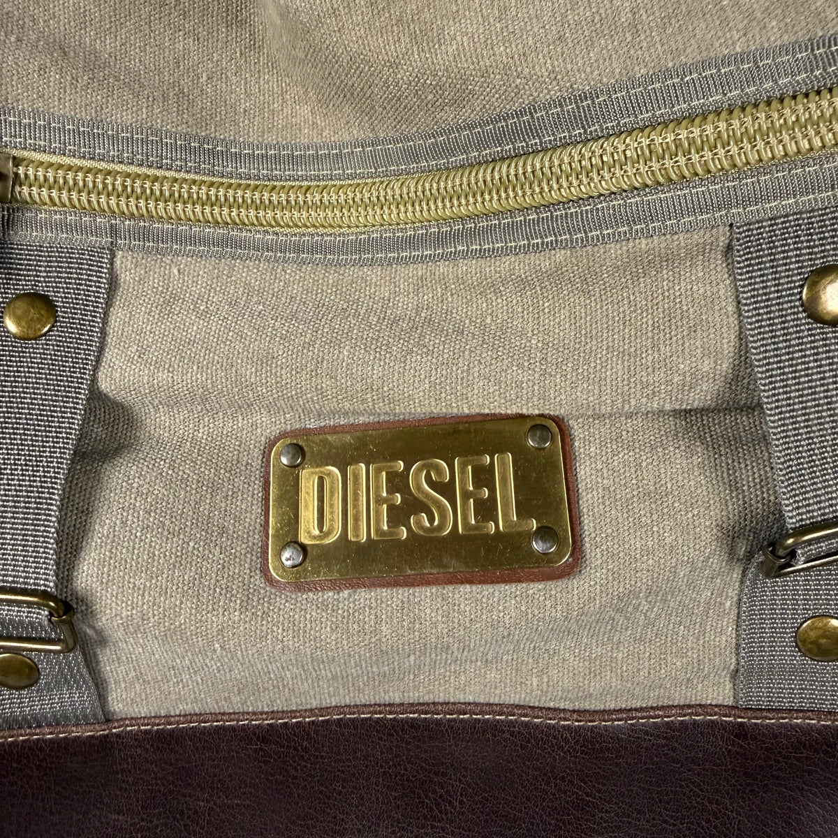Close-up of a Diesel brand brass plaque on a beige canvas bag with gray webbing straps and a gold zipper.