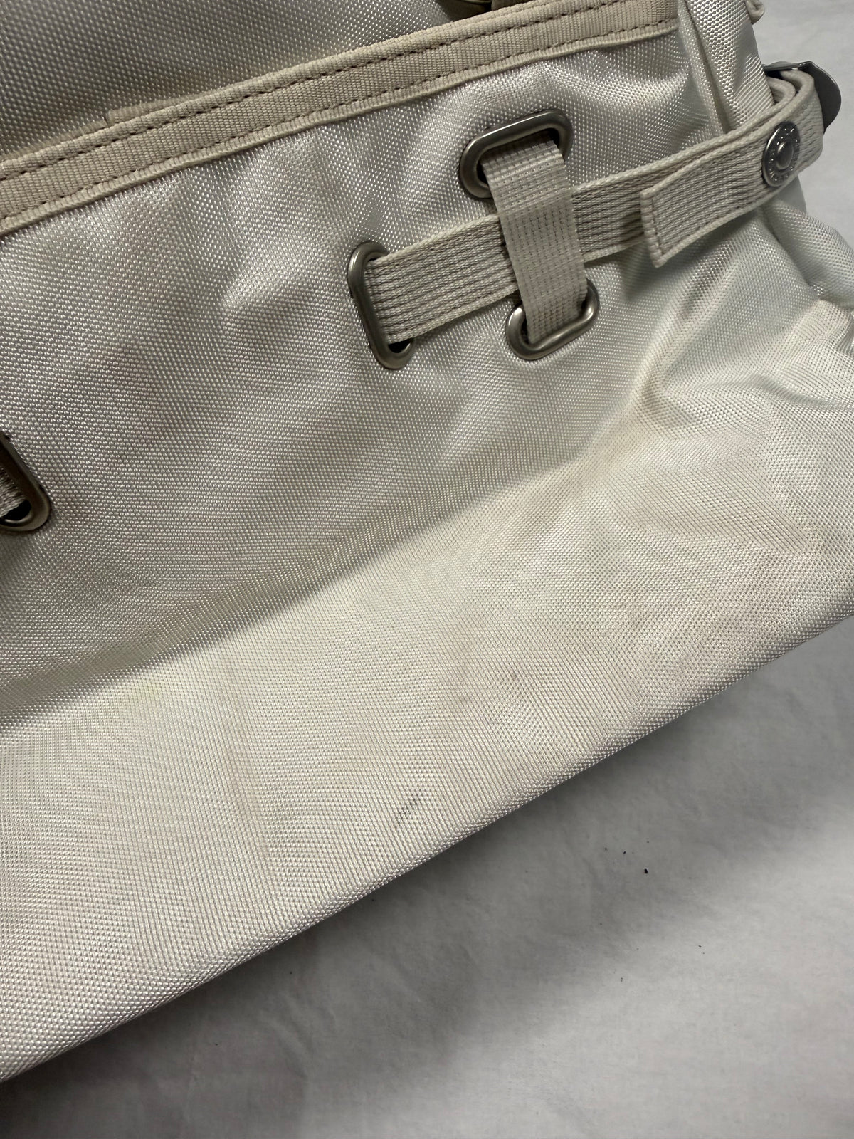 Close-up of a light beige canvas bag with a matching strap and silver metal grommets, resting on a light gray fabric surface.