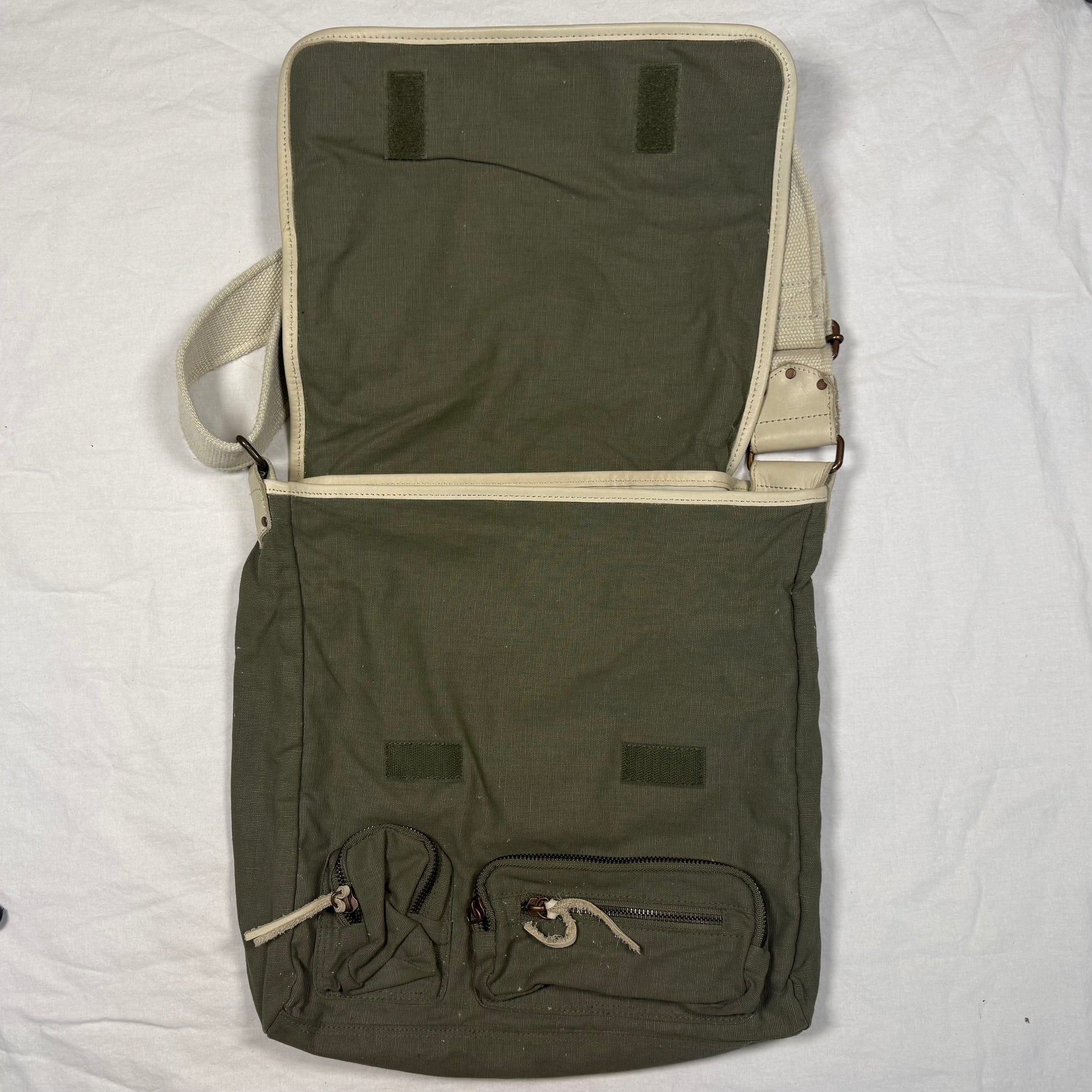 An olive green canvas messenger bag with beige trim, shown open to display its interior and multiple exterior pockets, laid flat on a white fabric background.
